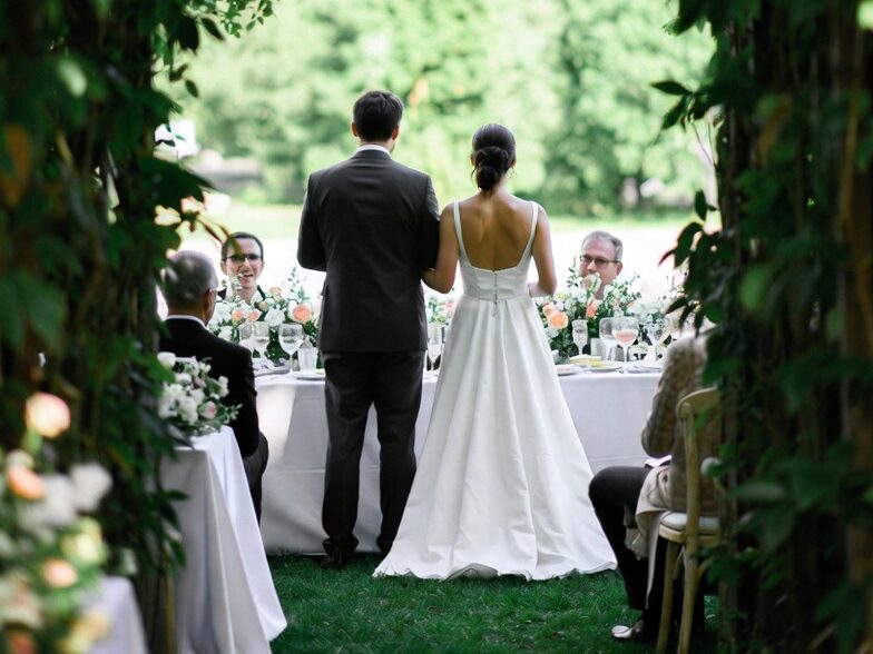Ein Brautpaar zieht flankiert von seinen sitzenden Gästen zum Hochzeitstisch.